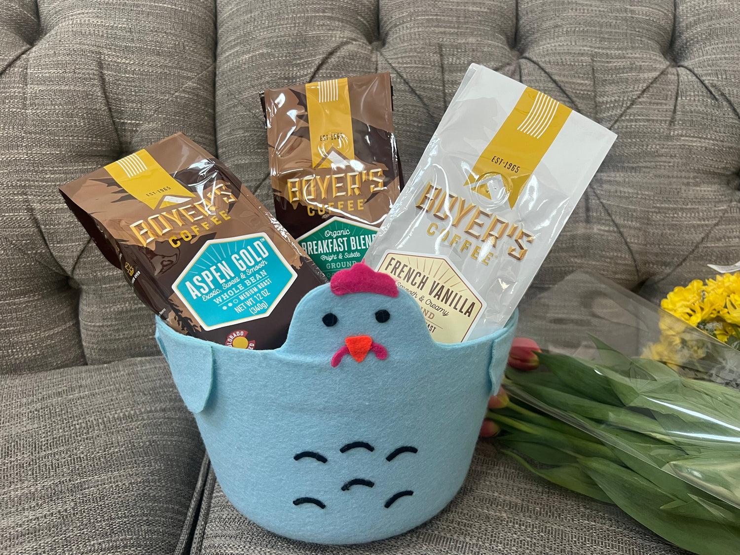 Blue chicken easter basket full of three bags of Boyer's Coffee with two bouquets of flowers sitting on a grey textured couch