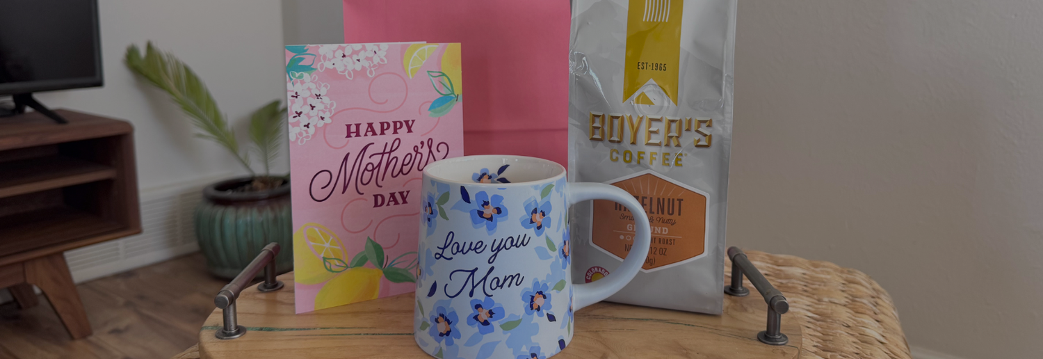 Bag of Hazelnut Coffee on a wooden tray, with a pink gift bag, mug that says "I love you mom" and mother's day card with a blurred living room background