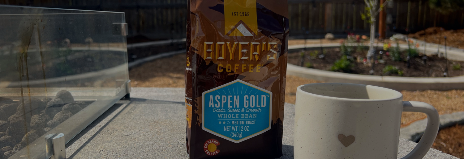 Boyer's Coffee Aspen Gold bag with a white mug on a reflective surface