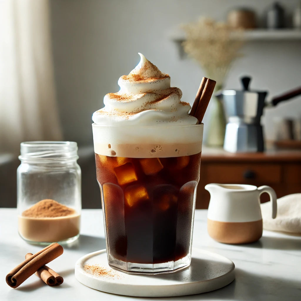 Iced coffee with whipped cream and a cinnamon stick on a table with coffee-making equipment in the background.