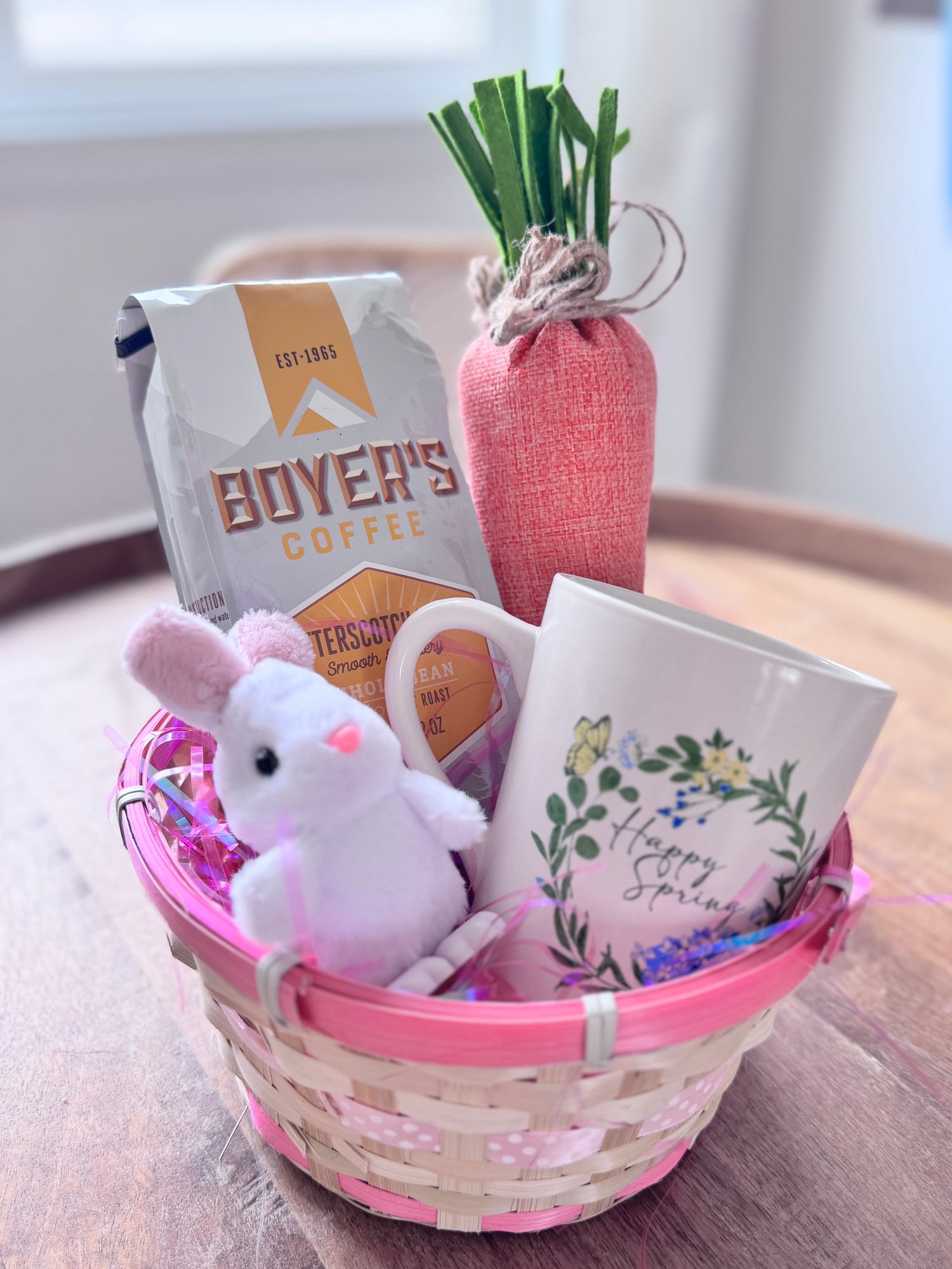 Easter Basket with pink ribbons full of a bag of Boyer's Coffee, a stuffed rabbit toy, a stuffed carrot toy and a mug that says "Happy Spring" sitting on a wooden table